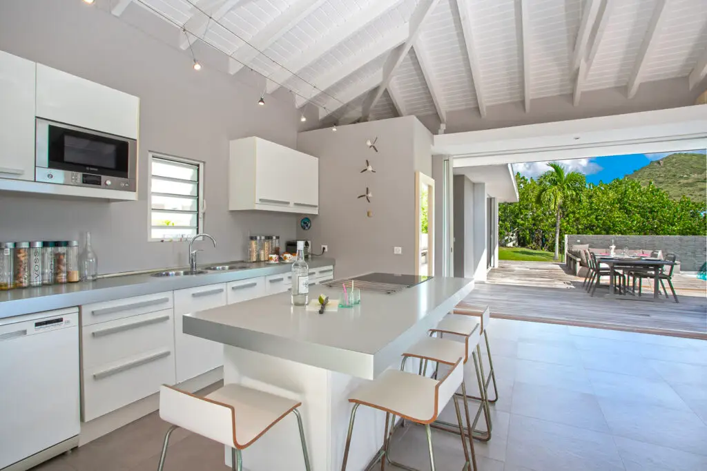 Modern open-air kitchen with sleek white cabinetry and island seating overlooking tropical gardens at La Villa, a nude clothing optional Caribbean resort