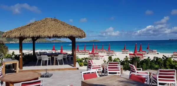 Thatched-roof beach cabana and red umbrellas at a beachfront restaurant on Orient Beach in Saint Martin, part of the nude-clothing-optional-caribbean resort area. Outdoor tables and lounge chairs face turquoise Caribbean waters under a bright blue sky.