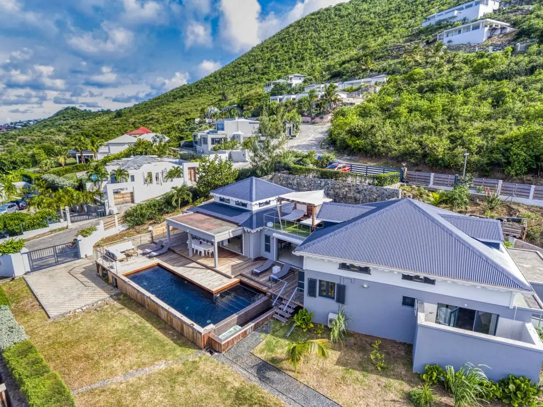 Aerial view of the nude-clothing-optional-caribbean Garden Villas resort in Saint Martin, showing modern gray-roofed villas surrounded by tropical hills. The image highlights a large wooden deck with a private swimming pool, outdoor lounge area, and lush green mountain backdrop.