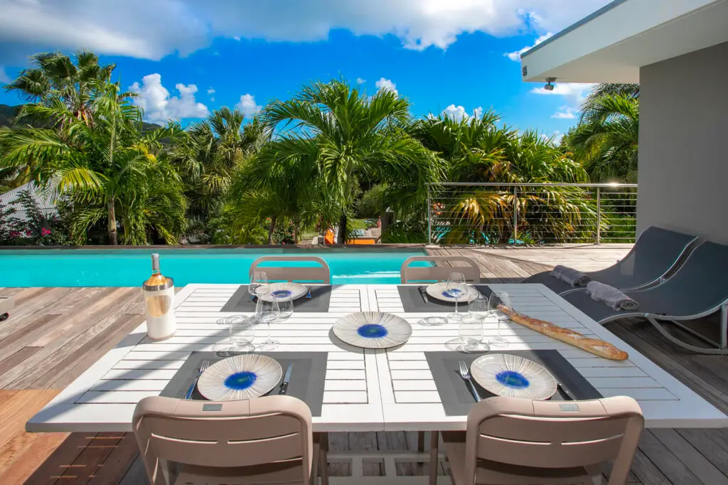 Elegant outdoor dining table set beside a private pool surrounded by palm trees at the nude-clothing-optional-caribbean Garden Villas resort in Saint Martin. Table set with wine, plates, and glassware under bright tropical skies, capturing a relaxed luxury atmosphere.