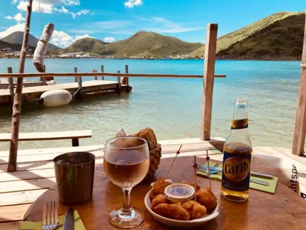 Table overlooking the turquoise bay in Saint Martin with a glass of wine, fried appetizers, and a Carib beer. Wooden pier and hills in the background capture the relaxed island dining atmosphere of the nude-clothing-optional-caribbean experience.