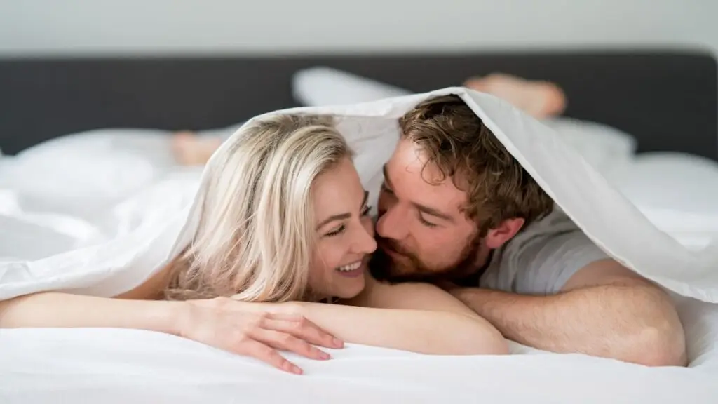 Smiling couple relaxing under a white sheet, representing intimacy and connection on a swinger cruise for lifestyle beginners