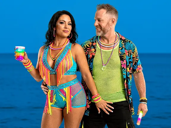 Couple dressed in colorful neon outfits with glow accessories and tropical drinks during Bliss Cruise swinger travel Glow fiesta theme night by the ocean